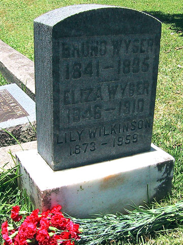 A dark headstone in Oahu Cemetery inscribed Bruno Wyser 1841-1895, Eliza Wyser 1846-1910, Lily Wilkinson 1873-1959, with red carnations laid at its base.