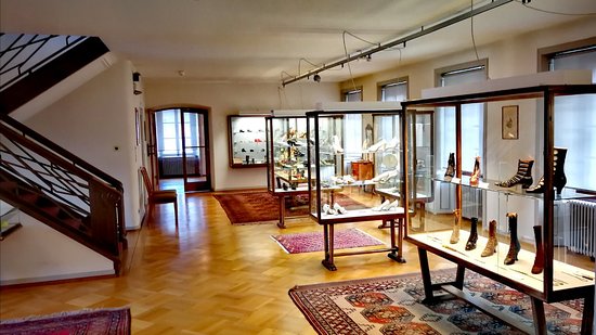 A museum room with parquet floors, oriental rugs and glass vitrines displaying antique boots and shoes.