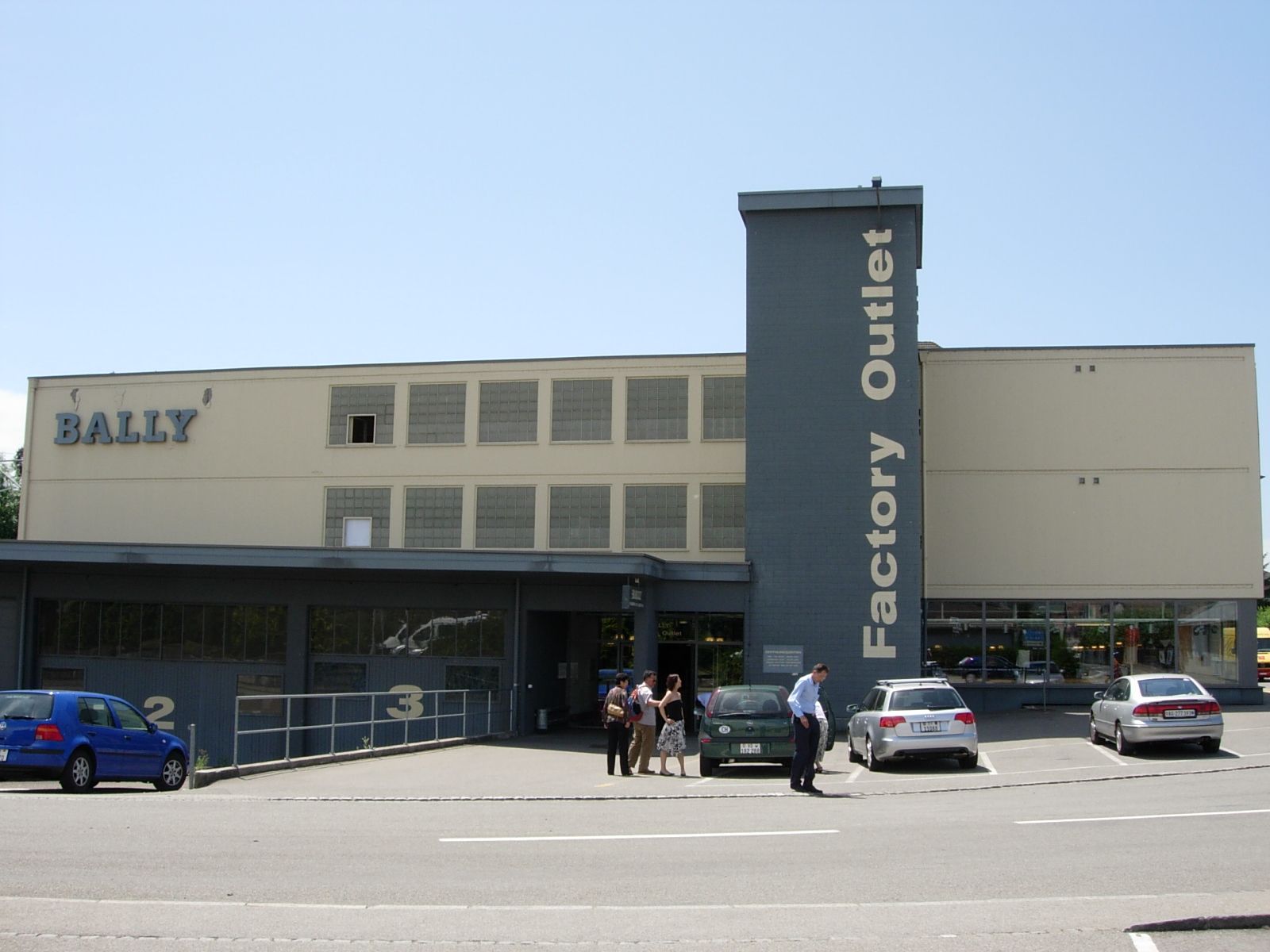 A low cream-and-grey industrial building with BALLY in large letters and a tower reading Factory Outlet.